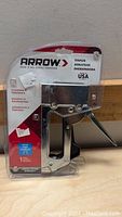 Photo of Arrow T50 stapler in clear plastic packaging, product visible with branding and usage information.