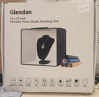 Box front showing brand, size 12x12 inches, portable photo studio shooting tent with icons indicating stepless dimming, high brightness, no strobe, assembly in seconds, multi-angle shooting, and included multi-color backdrops.