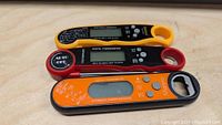 Photo showing three handheld digital thermometers in yellow, red, and black/orange colors with buttons and digital displays.