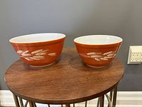 Two rust-colored Pyrex mixing bowls with white wheat designs placed side-by-side on a wooden surface against a grey wall background.