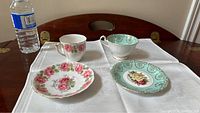 Two vintage Queen Anne fine china cup and saucer sets displayed on a table with a water bottle for scale. One set is white with pink roses and gold trim; the other set is turquoise with gold designs and rose motifs.