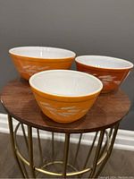 Three Pyrex Autumn Harvest Wheat pattern mixing bowls arranged on a round wooden table, showing the orange and rust alternating colors and wheat design on the exterior sides.