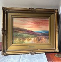 Framed watercolour landscape painting with golden ornate frame showing a hillside and colorful dramatic sky with a brass plaque beneath the painting.