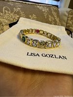 Gold Lisa Gozlan Adore bracelet resting on Lisa Gozlan branded pouch, showing gold-tone band with clear square stones and colored accent stones including a green round stone and a smiley face with black stones.
