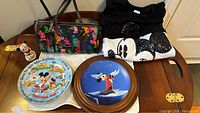Five-piece lot displayed on a table: colorful Dooney & Bourke handbag, XL black sequin Mickey sweatshirt, two Disney collectors plates, and Minnie skiing figurine.
