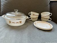 Photo of milk glass Anchor Hocking Fire King Wheat Pattern casserole with a clear glass lid and 4 Pyrex Butterfly Gold tea cups stacked two high and 4 matching saucers in front of the cups.