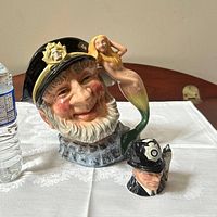 Lot showing The London Bobby figurine and Old Salt mermaid handle character jug on white tablecloth with water bottle for scale.
