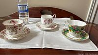 Three Royal Albert floral tea cups and matching saucers displayed on a white cloth on table with water bottle for scale.