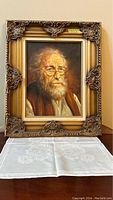 Framed oil painting of an elderly bearded man displayed on a table with a white cloth underneath, showing full frame and background.