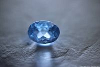 Oval cut aquamarine gemstone shown face-up on a textured surface with light blue color and multiple facets visible.
