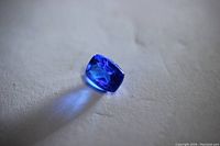 Close-up image of a single loose cushion cut blue Tanzanite stone placed on a light surface showing the stone's deep blue color and facets reflecting light.
