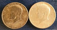 Obverse sides of two 1964 Kennedy half dollar coins, showing John F. Kennedy's profile and inscription details; one coin shows wear and discoloration, the other cleaner.