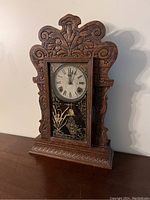 Front view of the antique wooden mantel clock showing the ornate carved wood frame, clock face with Roman numerals, and decorated glass door with gold painted bird and floral designs.