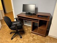 View of brown pressboard computer desk with black office chair and computer monitor.