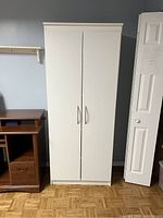 Tall white standalone cabinet closed, showing two doors with handles and crown molding top edge.