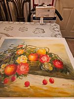 Photo showing the canvas painting laid flat on a table, displaying the detailed fruits and flowers in a basket with warm hues and artist's signature.