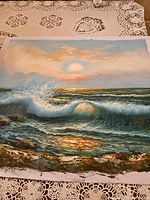 Canvas painting showing a detailed ocean water waves scene with a sunset sky in the background placed on a lace tablecloth.