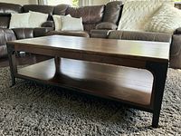 Full view of the solid wood rectangular coffee table with lower shelf, placed on a dark carpet in front of a sofa.