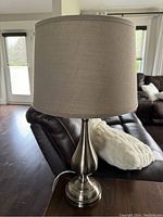 Photo showing one table lamp with a beige/taupe fabric drum shade and a brushed metal base, placed on a wooden surface with a leather couch in the background.