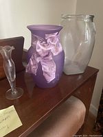 Three vases displayed on a wooden surface: clear oval vase, purple vase with satin bow, and small clear fluted vase.