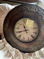 Front view of a large 28-inch round wall clock displaying Roman numerals and ornate gold detailing on a brown frame, lying on beige fabric surface.