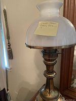 Early American style brass lamp with white opaque glass shade shown standing on a surface. The lamp is approximately 19 inches high with a polished brass base and fitting, topped by a white glass shade with flared edge.