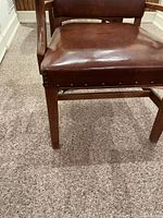 Close-up of the brown leather seat showing nailhead trim and wooden chair leg on carpeted floor.