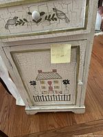 Front view of the small side table showing the drawer with bird design and lower cabinet with house and flower motif crackle paint decoration.