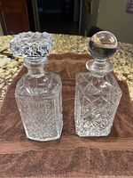 Two crystal whiskey decanters shown side by side on a brown cloth on countertop, displaying their unique stopper shapes and cut glass designs.