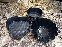 Initial overview photo showing multiple black springform pans including round and heart-shaped, and a flower-shaped brioche pan on granite countertop.