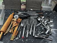 Photo showing large wooden rolling pins, wooden spoons, whisks, knives, ladles, and tongs neatly laid out on table