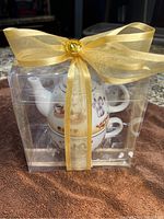 Clear plastic gift box containing teapot and stacked cups, wrapped with yellow ribbon and decorative gem, photo shows front view of the set