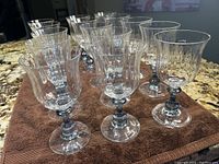 Front and side perspective of 15 clear crystal wine goblets with blue stems on a brown cloth.