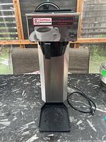 Front view of industrial coffee maker with power cord visible, placed on black table.