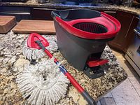 Vileda mop bucket in gray and red with mop handle and two mop heads on granite countertop.