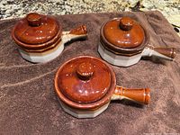 Three pottery French onion soup bowls arranged on a towel, showing the brown glossy lids and handles, beige bases, all in good condition.