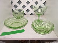 Four pieces of green uranium glass displayed together on a white shelf. The set includes a plate, a goblet, a small bowl, and a dessert glass. The pieces are translucent green and glow under black light.