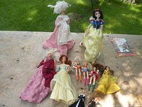 Ten Barbie dolls displayed on a wooden surface outside. Two large dolls, one in yellow satin and one in pink crocheted dress, with eight smaller dolls in various outfits.