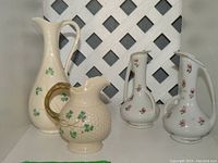 All four small jugs arranged on white lattice background showing two Belleek jugs with shamrocks on the left and two Occupied Japan jugs with floral decorations on the right.