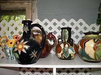 Photo showing five ceramic jugs with colorful floral and abstract painted styles placed on a white shelf against a gray background