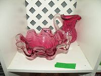 Cranberry glass ruffled edge bowl with clear accents and swirl design alongside cranberry glass pitcher