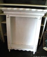 Front view of white farmhouse style wall shelf cupboard showing decorative top molding, turned spindle legs, and scalloped bottom edge.