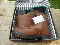 Side view of brown Kozi boots in striped box on wooden surface showing lace-up front, side zipper, and rubber sole.