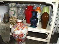 Lot of various vases on a white wicker shelf, showing different colors, shapes and sizes of the vases.