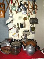 Photo of lot of vintage kitchen utensils hanging on wooden expandable racks and pegboard wall including kettles, whisks, metal mugs, egg beaters, scrubbers, and a small toaster