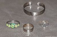 Four silver jewelry pieces on surface: baby cuff, three silver rings including one with green stones and one with anchor design.