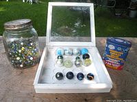 Full view of lot including large glass jar filled with marbles on left, white display case with 15 marbles in center, and Marvelous Marbles box on right outdoors on a table with grass background.