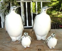 Front view of 2 large and 2 small white and gold penguin figurines on a wooden surface in outdoor setting.