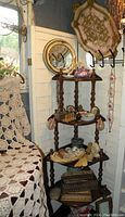 Full view of tiered wooden knickknack shelf holding various vintage vanity items including porcelain box, shell dishes, and small decorative items. Large decorative metal tray and round gold mirror mounted on wall behind.