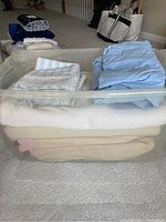 Photo of clear plastic storage bin packed with assorted bed linens including blankets, quilts, sheets in various pastel and neutral colors folded inside